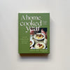 A green cookbook called 'A Home Cooked Year' by Annie Mae Herring photographed on a neutral backdrop.