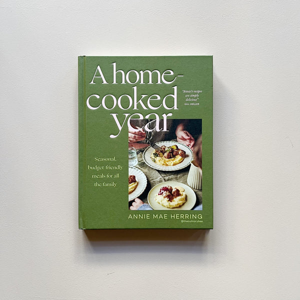 A green cookbook called 'A Home Cooked Year' by Annie Mae Herring photographed on a neutral backdrop.