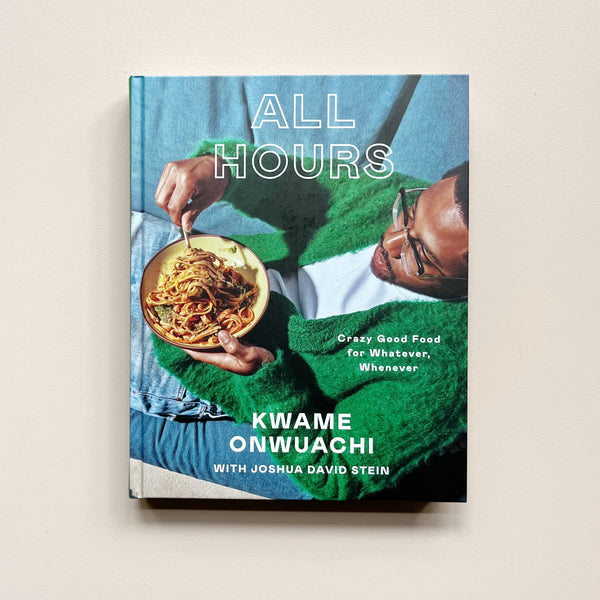 A hardcover cookbook titled 'All Hours: Crazy Good Food for Whatever, Whenever' by Kwame Onwuachi with Joshua David Stein. The cover features a man in a green sweater and jeans eating a bowl of noodles on a blue couch. The book is sitting on a neutral background.