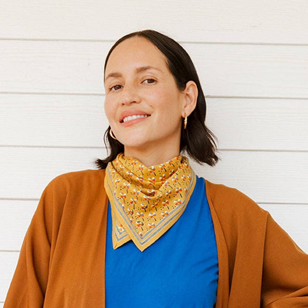 Woman wearing a brown coat over a blue outfit with a yellow bandana, standing against a white wall.