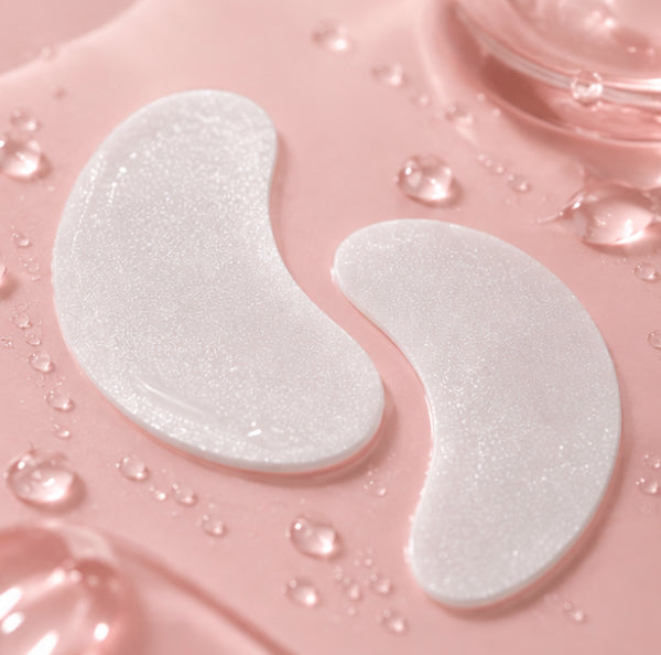 Two white crescent-shaped under eye patches resting on a pink surface with water droplets.