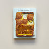 A cookbook called "Cheese Please" by Sue Quinn showing a cripsy oven-roasted dish on  a white cover. The book sits against a neutral backdrop.