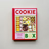 A baking book called 'Cookie Box' in bright red, pink and pale green with an image of a box of various cookies. The book sits against a neutral backdrop.
