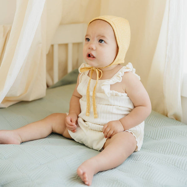 Baby in a yellow bonnet sitting on a crib with beige curtains in the background