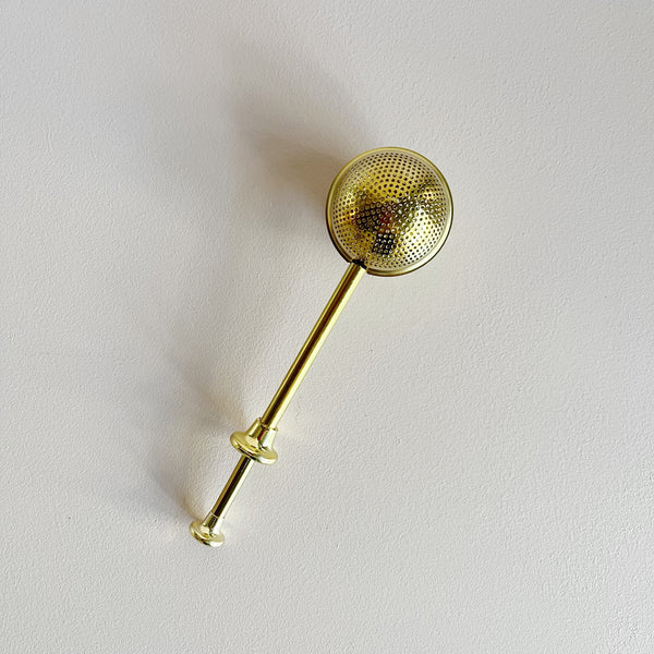 Gold toned stainless steel tea infuser on a neutral background.