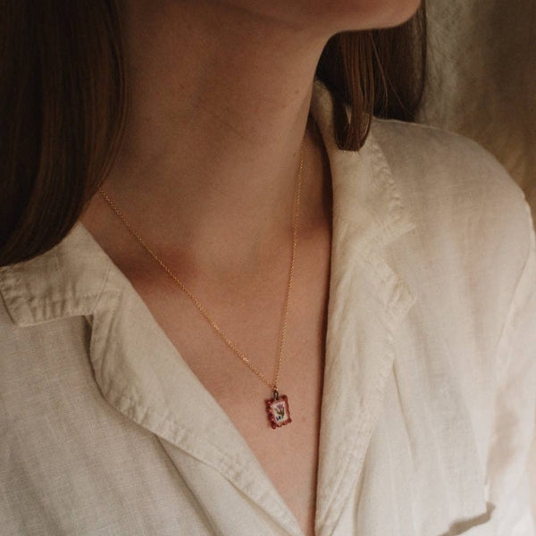 Person wearing a light-colored shirt and a delicate necklace with a small postage stamp pendant.