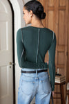 A woman shown from the back wearing a fitted long-sleeve top in dark forest green with contrast stitched side seams and a vertical seam down the back, paired with light blue jeans.