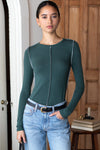 A woman modeling a dark teal green long-sleeve knit top with a central contrast stitched seam detail, paired with light-wash blue jeans and a black belt, standing in front of a wooden door.