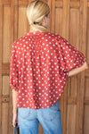 A woman viewed from behind wearing a red short-sleeve blouse with a white block printed diamond pattern, paired with blue jeans, standing in front of a wooden door.