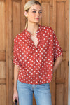 A woman wearing a rust-red short-sleeve button-up blouse with a white block printed diamond pattern, paired with light-wash blue jeans, standing in front of a rustic wooden door.