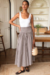 A woman stands in a bright indoor room wearing a sleeveless white top and a brown-and-white gingham maxi skirt, holding a woven straw handbag.