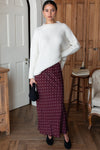 A woman stands in a bedroom wearing a white sweater over a rich tawny red and white polka dot bias cut maxi skirt.