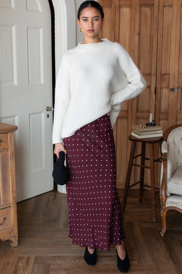 A woman stands in a bedroom wearing a white sweater over a rich tawny red and white polka dot bias cut maxi skirt.