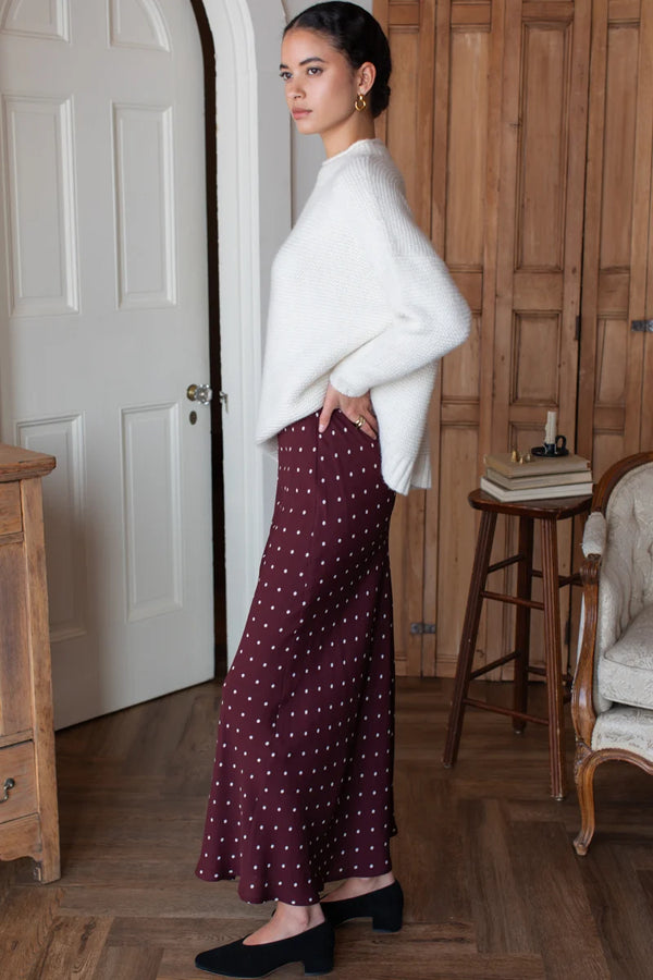 A woman stands to the side in a natural lit bedroom, wearing a white sweater and black shoes with a rich tawny red and white polka dot bias cut maxi skirt.