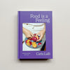 The book 'Food is a Feeling' by Carla Lalli, featuring a solid purple cover with white text and a central photograph of hands peeling fruit from an apple, displayed against a neutral light gray background.