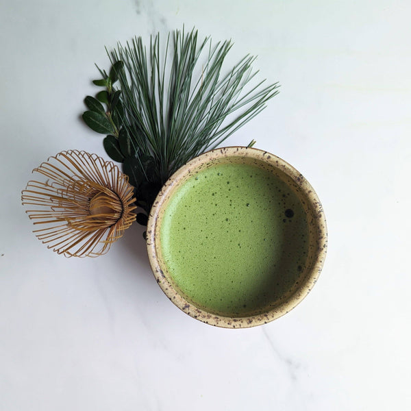 Ceramic bowl with green matcha, a whisk, and greenery on a white background