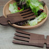 Wooden salad servers on a wooden table with a plate of salad.