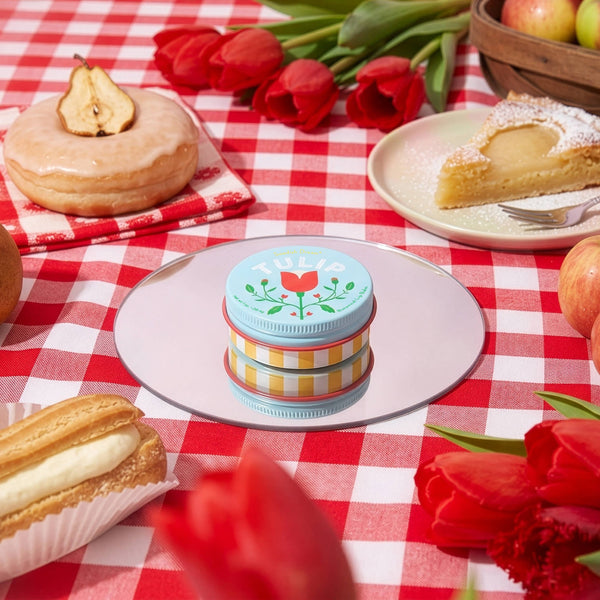 A tin of Tulip botanical lip balm on a round mirror surrounded by fruits, desserts, and red tulips against a red and white gingham picnic blanket.