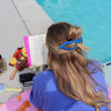 Person sitting by a pool with a book and snacks, wearing a blue and white polka dot hair claw.