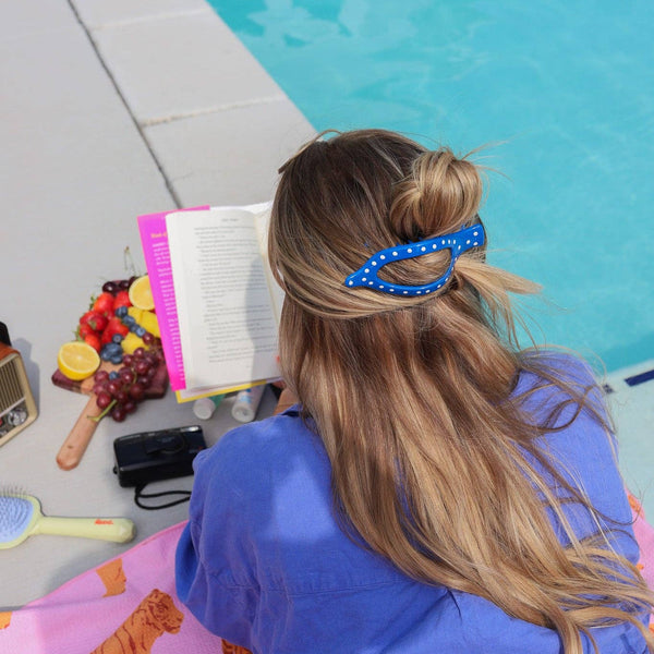 Person sitting by a pool with a book and snacks, wearing a blue and white polka dot hair claw.