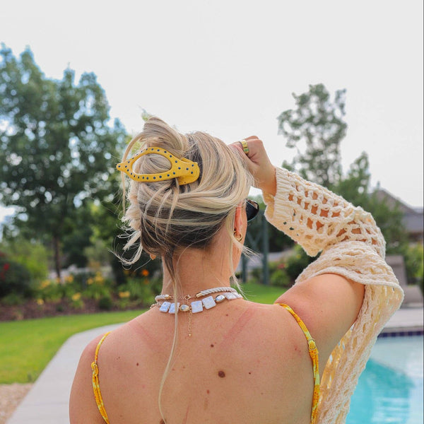 Person wearing a yellow and blue polka dot hair claw in front of an outdoor pool and trees.