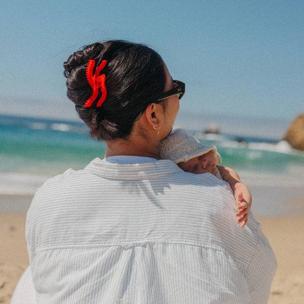 Woman holding a baby on a beach with clear blue sky and ocean. She is wearing a bright red hair claw. 