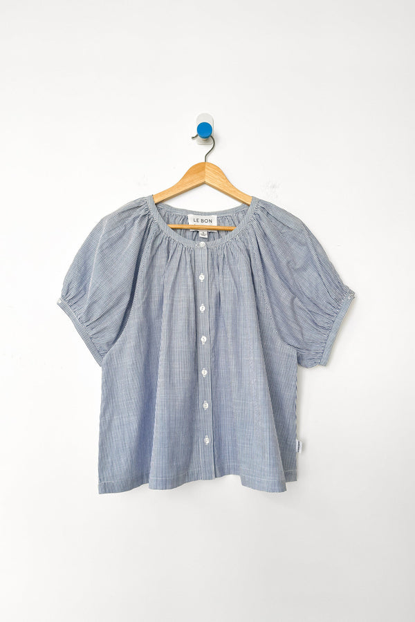 A blue and white striped cotton short sleeve blouse on a wood hanger against a white wall.