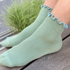 Person wearing green socks with blue ruffle edge on a wood surface with greenery in the background.