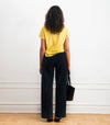 A woman stands with her back to the camera wearing a bright yellow short-sleeve t-shirt and dark wide-leg jeans, holding a black bucket bag against a white wall.