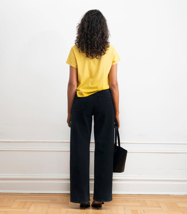 A woman stands with her back to the camera wearing a bright yellow short-sleeve t-shirt and dark wide-leg jeans, holding a black bucket bag against a white wall.