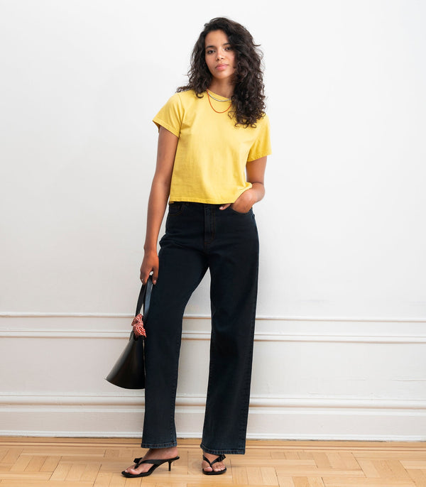 Woman wearing a yellow top and black high rise wide leg jeans holding a black handbag against a white wall.