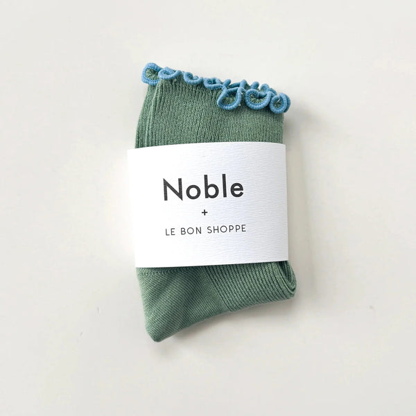 Green socks with blue ruffle edge in white paper branded packaging.