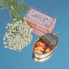 A tin can viewed from the top, filled with pickled mussels, alongside a floral decoration and a Siesta Co box. The mirror background is a sky blue color. 