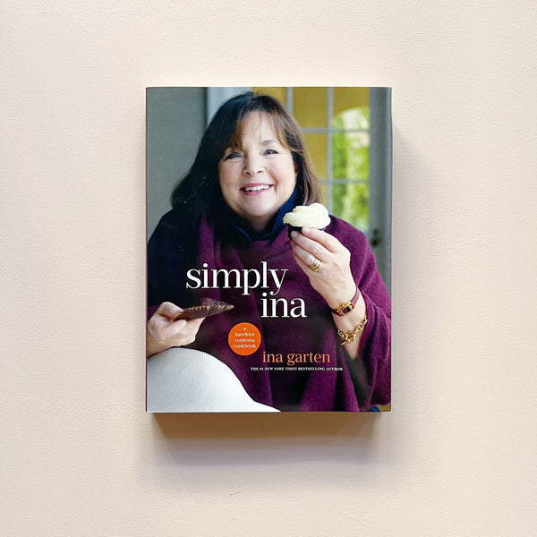 A cookbook called 'Simply Ina' with a photo on the cover of Ina Garten holding a cupcake sitting in a home environment.