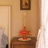 Candle on a red horse shaped holder on a wooden table with a framed picture on the wall.