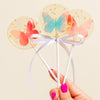 Three clear and gold flecked lollipops with colorful butterflies held by a hand with pink nail polish against a beige background.