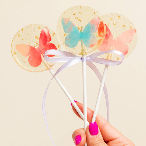 Three clear and gold flecked lollipops with colorful butterflies held by a hand with pink nail polish against a beige background.