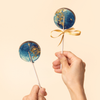 Two blue lollipops with gold accents and a hand holding one, on a beige background. Brand 'Sweet Caroline Confections' visible.