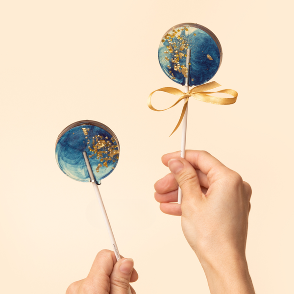 Two blue lollipops with gold accents and a hand holding one, on a beige background. Brand 'Sweet Caroline Confections' visible.