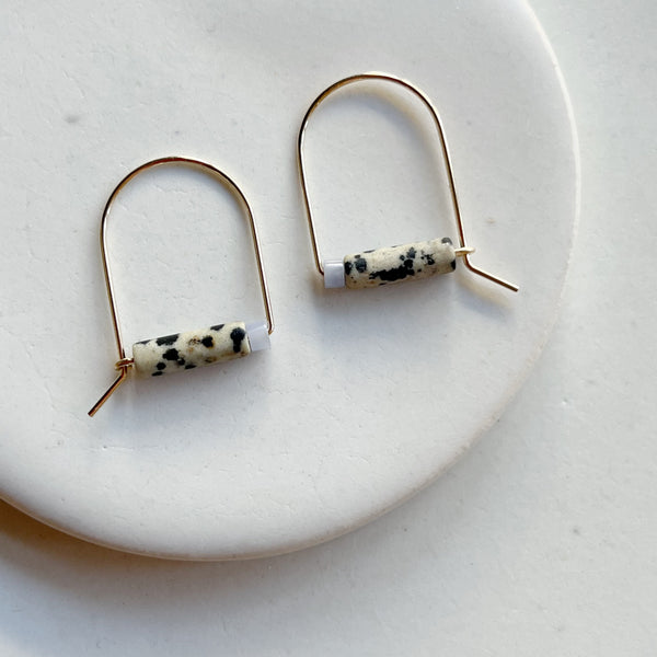 Pair of gold arch earrings with dalmation jasper cylindrical beads on a white background.