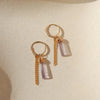 Gold hoop earrings with amethyst stone and gold chain drop on neutral background.