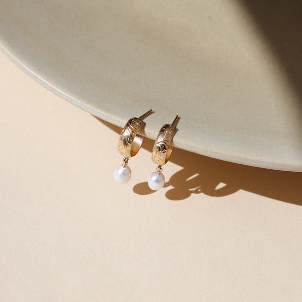 Gold stamped hoops with pearl charm dangles on a neutral ceramic backdrop.
