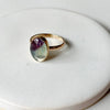 A gold ring with a natural purple and green fluorite oval stone on a white ceramic backdrop. 