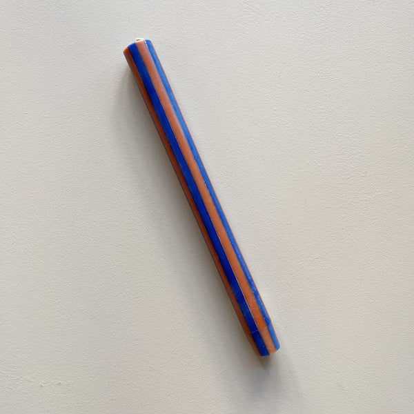 A taper candle in a vertical striped navy and orange design against a neutral backdrop.