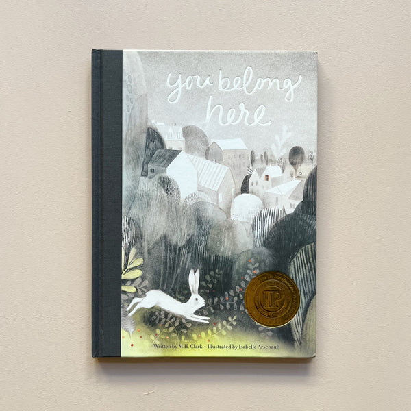 An illustrated children's book called 'You Belong Here' featuring a greyscale illustration of a town with a white rabbit in the foreground. The book sits against a neutral backdrop.