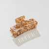 A rectangular claw hair clip with a glossy tortoiseshell pattern in shades of brown, orange, and cream, displayed on a clean white surface with a soft reflection.