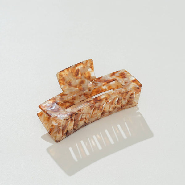 A rectangular claw hair clip with a glossy tortoiseshell pattern in shades of brown, orange, and cream, displayed on a clean white surface with a soft reflection.