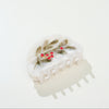A curved hair claw in iridescent white with a green mistletoe sprig accented by red bejeweled berries, photographed against a white backdrop with a soft shadow.
