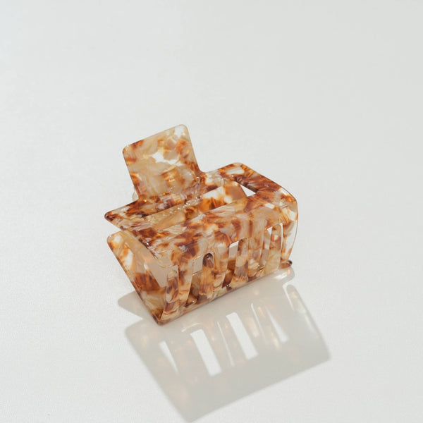 A square translucent tortoiseshell hair claw clip with a warm brown and cream marbled pattern, shown on a clean white reflective surface.