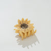 A small sunflower-shaped hair claw clip in a warm pale yellow tone, with a dark brown patterned center and thin gold metallic rays, photographed on a clean white background.